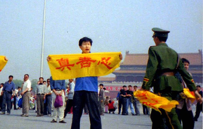Un policier chinois s'approche d'un pratiquant de Falun Gong sur la place Tiananmen à Pékin alors qu'il tient une bannière avec les caractères chinois signifiant « Authenticité, Bienveillance et Tolérance », les principes fondamentaux du Falun Gong. (via Minghui.org)